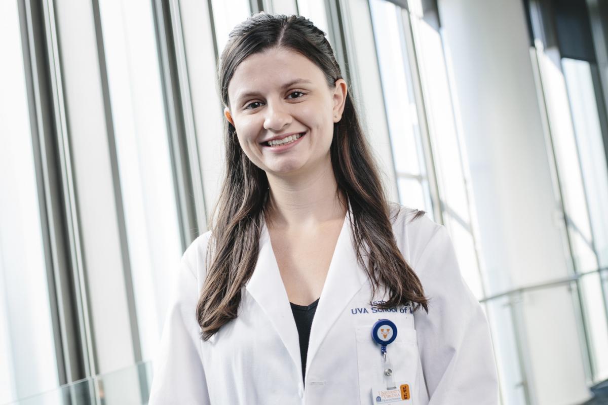 Isabelle Gill wears a white coat and stands in front of windows in a UVA Health building.